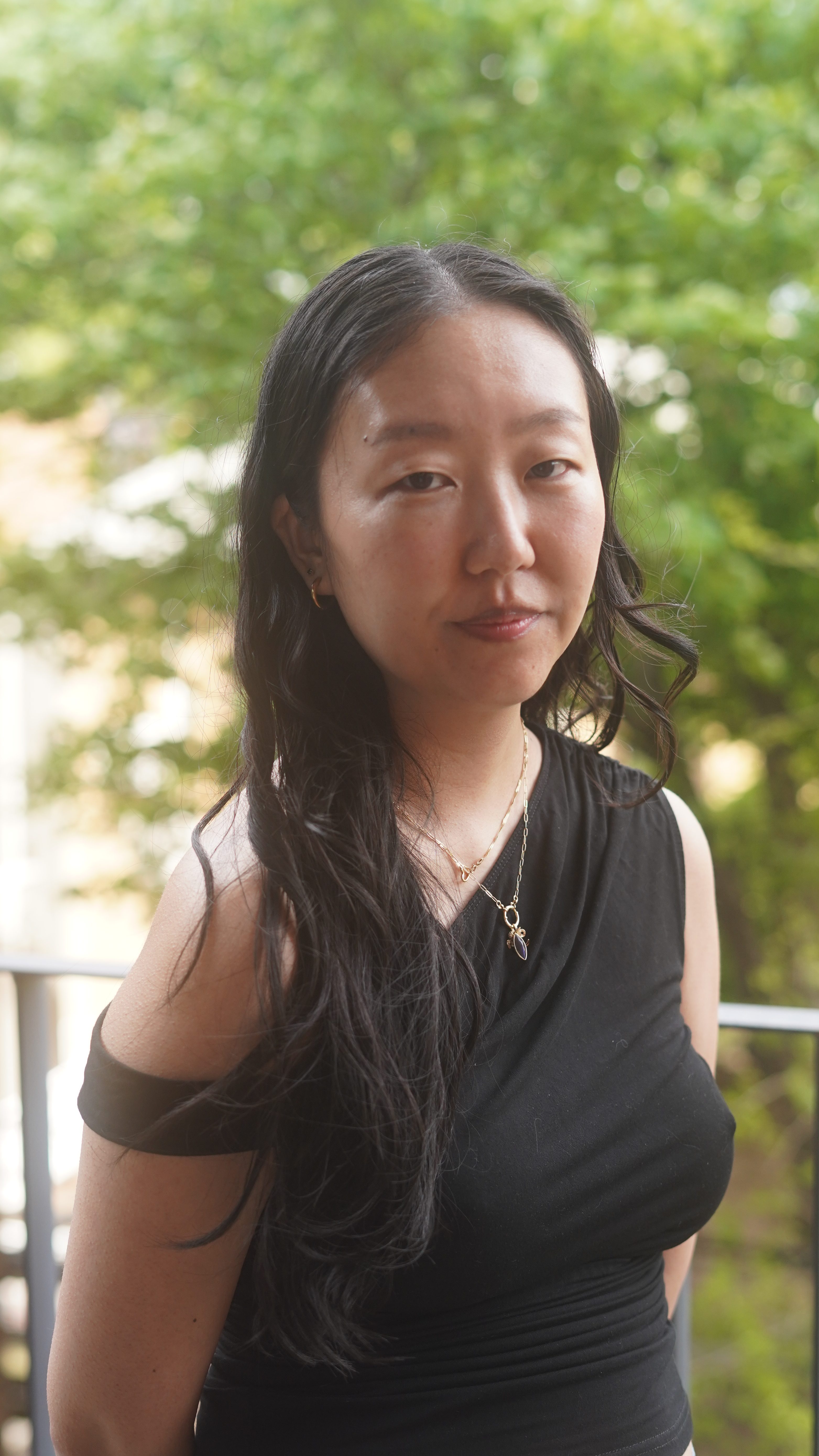 Christina is a Korean person with long dark wavy hair. She is looking into the camera steadily, and is wearing a black asymmetrical shirt. 